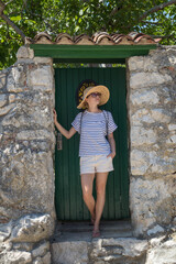 Beautiful young female tourist woman wearing sun hat, standing and relaxing in shade in front of vinatage wooden door in old Mediterranean town while sightseeing on hot summer day.