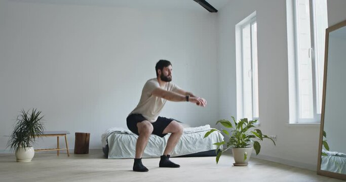 Young bearded man doing squats exercises at home