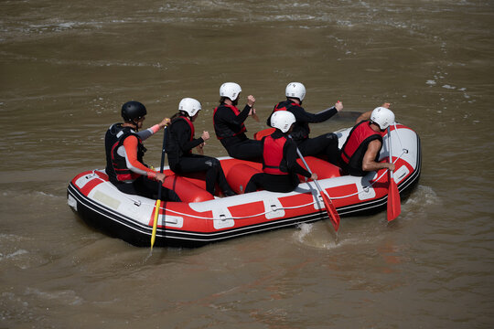 Rafting Team Members And Instructor Rowing In Their Boat Down The Murky River During A Summer Day