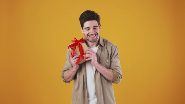Young bunet man is catching gift box tied with red ribbon. Smiling and looking overjoyed while posing against orange studio background. Close up