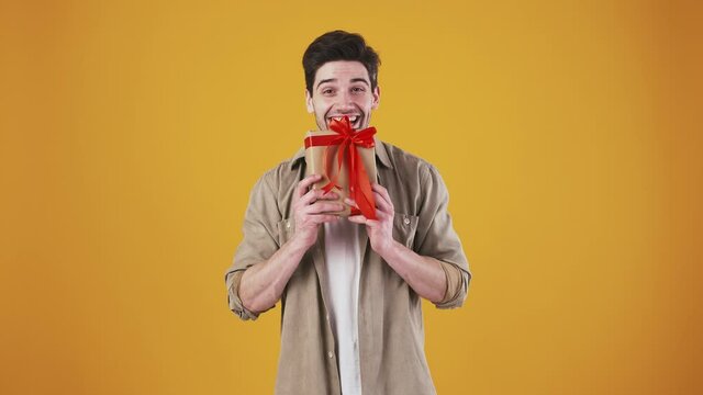 Male is smiling, shaking gift box trying to guess what is there inside. Rejoicing while posing against orange studio background. Birthday