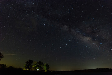Fototapeta premium Trees lit up by campers on lake shore under the Milky Way