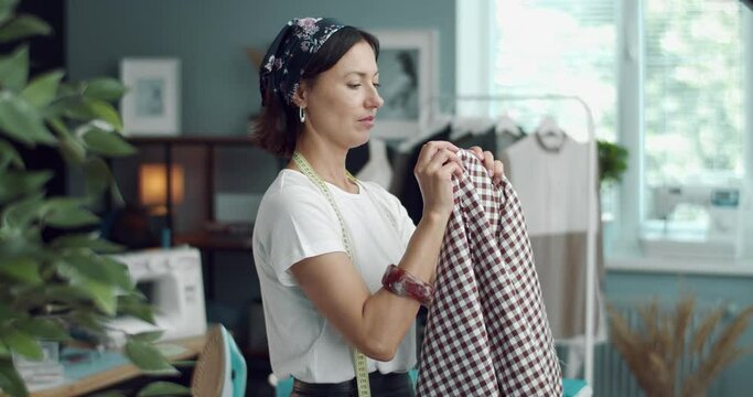 Side view of fashion designer estimating checkered fabric for new clothing collection. Mature woman with brown hair testing textile at modern studio.