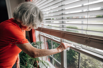 An elderly woman cleaning and sanitizing her window sill and blinds in her home
