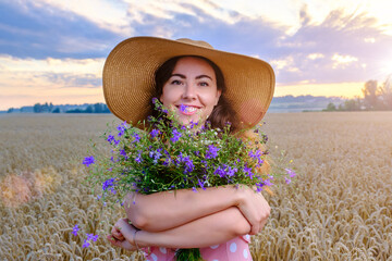 Pretty young woman in straw hat holds a wild flower bouquet in ripe wheat field at sunrise and happily smiling.  © Serhii