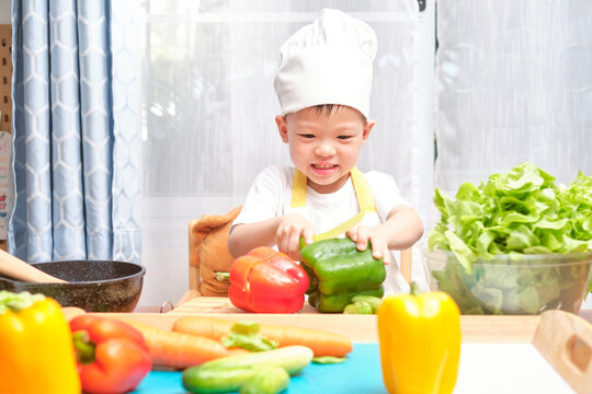 Cute Happy Smiling Asian Little Boy Child Wearing Chef Hat And Apron Having Fun Preparing, Cooking Healthy Food, Kid Use Safe Kid Friendly Plastic Kitchen Knife Cutting Vegetables In Kitchen At Home