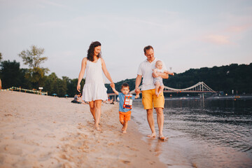 family vacation in summer. Young Caucasian family foot walking barefoot sandy beach, shore river water. Dad mom holding hands two children, brothers. Big friendly family with two children near lake