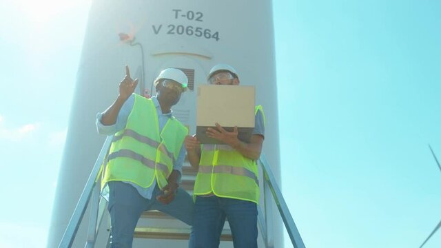 Engineers men in uniform discuss use laptop stand near wind turbines ecological energy industry power windmill field worker renewable slow motion
