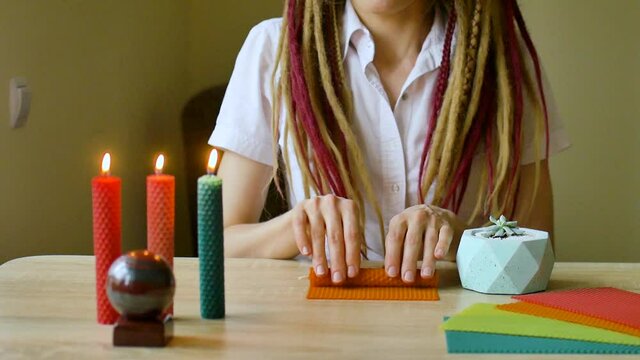 Young modern girl with dreadlocks in white shirt is performing a workshop of making beeswax candles sitting in front the table with stone and plants on it