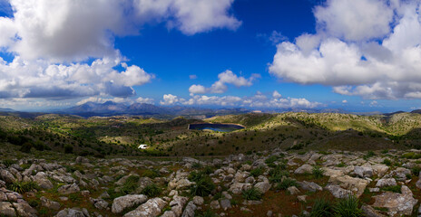 Fototapeta na wymiar Wide panoramic view of the island of Crete in Greece. Landscape with a view of a water quarry, mountains and blue sky in clouds, with rocks and grass bushes in the foreground