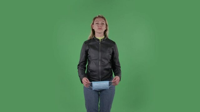 Portrait of beautiful young woman puts on medical mask. Blonde with loose hair in a black jacket and jeans on a green screen in the studio. Health Protection Corona Virus Concept. Slow motion.