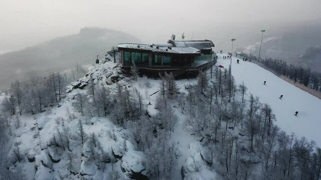 Aerial view of the ski resort with snowy mountain slopes and winter trees. Stock footage. Upper station of cableway and a group of people going skiing and snowboarding down the hill.