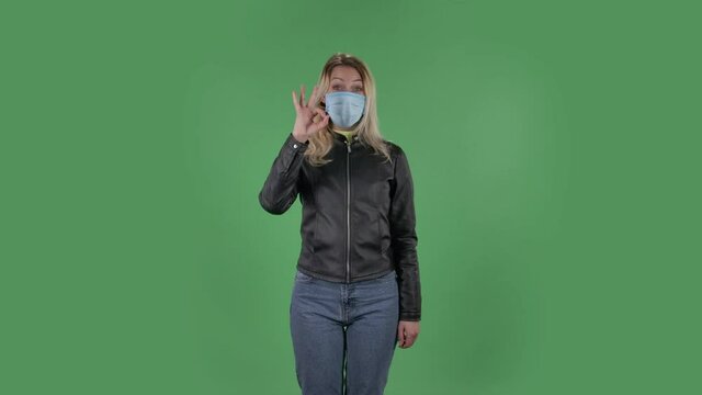 Portrait of beautiful young woman in medical mask is looking at camera and joyfully making sign ok. Blonde with loose hair in a black jacket and jeans on a green screen in the studio. Health