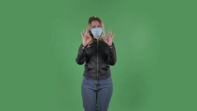 Portrait of beautiful young woman in medical protective face mask looking at camera and showing thumbs up and making sign ok with both hands. Blonde with loose hair in a black jacket and jeans on a