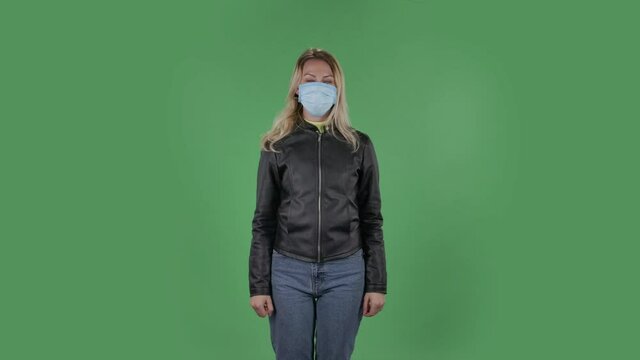 Portrait of beautiful young woman in medical mask is looking at camera and pointing up fingers. Blonde with loose hair in a black jacket and jeans on a green screen in the studio. Health Protection