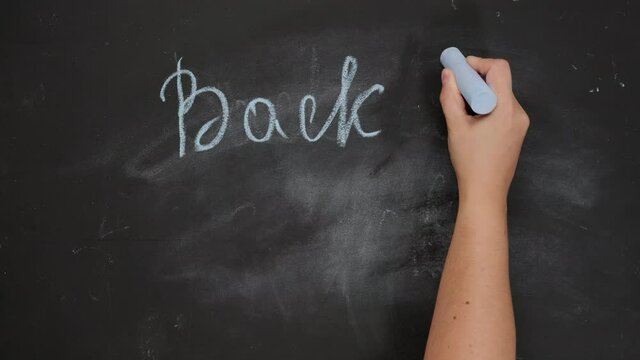 female hand holds blue chalk and writes back to school on black chalk board, start of the school year