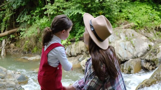 Loving young mother kisses cute little daughter standing near amazing small waterfall mountain river on background green forest. Concept of affection, adventure, family bonds care and protection