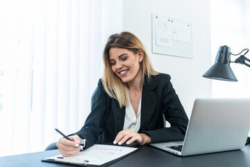 Smiling female entrepreneur in suit sitting in office and writing on paper.