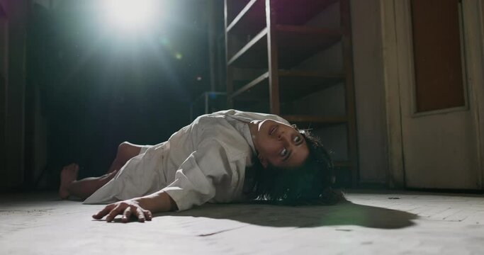 Exhorcist girl in white shirt convulsing on old wooden floor. Scary female demon awakening in haunted hallway - halloween costume, horror movie 4k footage