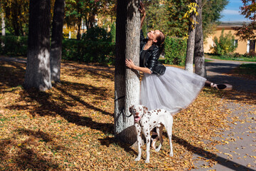 Ballerina with Dalmatian dog in the Park. Woman ballerina in a white ballet skirt and black leather jacket dancing in pointe shoes in autumn park with her spotty dalmatian dog.