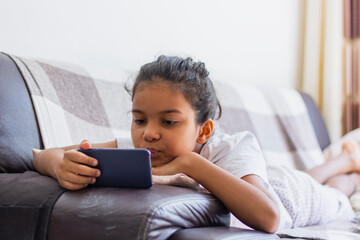 Brazilian kid Using Mobile Phone Watching Cartoons At Home, Lying On Sofa Relaxing On Weekend. Kid And Gadget Concept