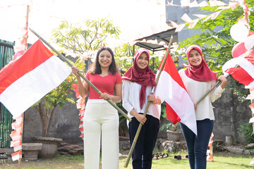 three asian girls in red and white shirts and headbands stand holding bamboo sticks with Indonesian flags to celebrate Indonesian Independence Day