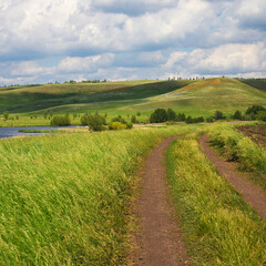 A country road among fields with grasses, near a lake and hills. Summer natural landscape with cloudy sky.
