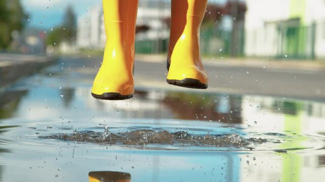 SLOW MOTION, LOW ANGLE, CLOSE UP, DOF: Unrecognizable young woman wearing jeans and yellow rain boots jumps in the glassy puddle on the empty sidewalk after a long spring rainstorm. Carefree girl.