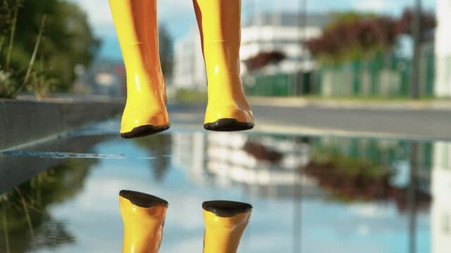 SLOW MOTION, LOW ANGLE, CLOSE UP, DOF: Unrecognizable girl in brand new yellow rubber boots jumps into the glassy puddle on the sidewalk. Carefree young woman in rain boots jumps into the big puddle.