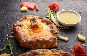 Two fresh and delicious khachapuri of weed grain flour and cottage cheese, egg, sulugini cheese, vegetables, oil, cutlery on a dark background.