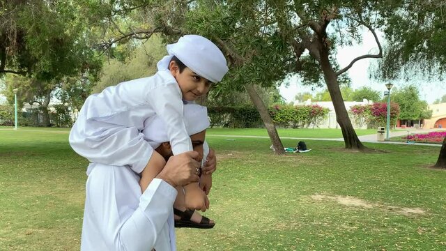 Happy Arab Middle Eastern Family at Park. Arabic father and son spending time together wearing traditional UAE Middle East Kandura dish dash. Emirati dad carries his son on his shoulders