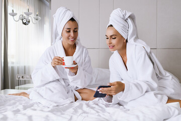 Two females sit at luxury appartment hotel bed at morning with cup, wear bathrobes and towels, look at smartphone, online networking, shopping, chatting, ladies friendship and leisure
