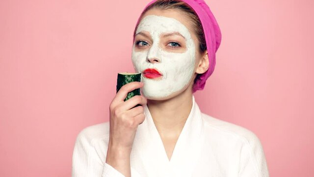 Girl with a mask on her face eating cucumber on pink background. Woman wearing face mask. Face care, facial beauty concept.