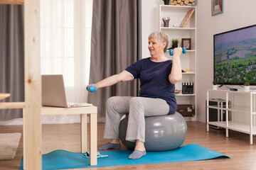 Pensioner learning online biceps training sitting on swiss ball. Old person pensioner online internet exercise training at home sport activity with dumbbell, resistance band, balance ball at elderly