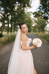 Gorgeous bride in elegant wedding dress stand in linden alley with bouquet and posing to photographer