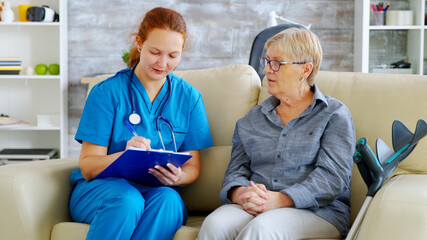Obraz premium Female doctor with red hair taking notes on clipboard while talking with senior woman on the couch in nursing home