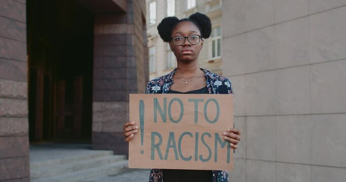 Portrait of african american girl looking to camera while holding placard no to racism. Woman with nose ring protesting against racial inequality at city street. Concept of human rights