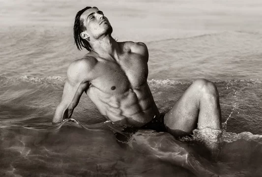 Sexy fit male model with hot strong body lying in the water at the beach.