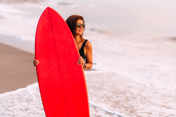 Beautiful sexy surfer girl on the beach at sunset. Beautiful girl with a surfboard. Attractive sexy young woman in black swimsuit is standing on beach with red surfboard in hands. Copy space
