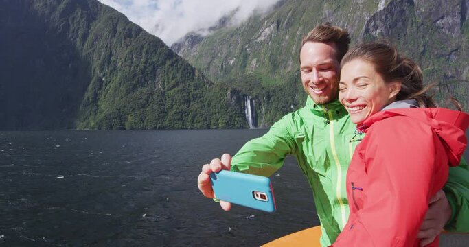 Happy couple on travel vacation taking phone selfie using smartphone on cruise ship, Milford Sound, Fiordland National Park, New Zealand. Multicultural couple smilng having fun laughing SLOW MOTION.