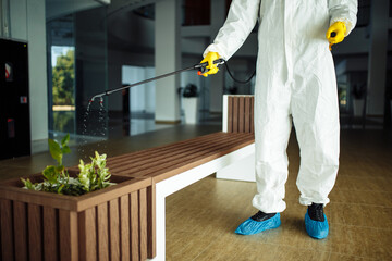 A man wearing protective suit is disinfecting a bench in an empty shopping mall with sanitizing spray. Cleaning up the public place to prevent covid spread. Healthcare precautions and safety concept.