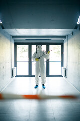 Fototapeta premium A man wearing protective disinfection suit and a spray stands in front of a glass doors under the staircases. Worker cleaning up the business center. Healthcare, covid-19 concept.