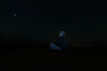 A lonely man sitting on the desert sand looking at the starry sky, between dunes in the desert of Erg Chebbi