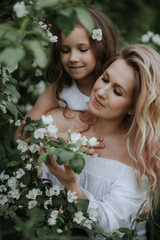 Happy mother and daughter in white clothes and with flowers in their hair look at the white flowers and smile.Daughter gently embraces her mother against the background of greenery