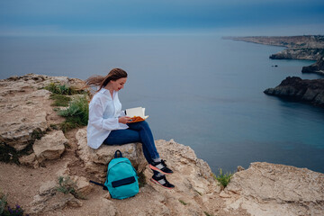 Fototapeta premium Attractive woman is sitting on the edge of the mountain with a sea view, reading a book.