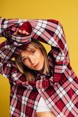 Stylish trendy young woman posing on camera with hands above head. Girl in red checkered shirt alone in studio. Clothing advertisement. Isolated over yellow background.
