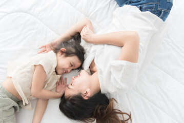 Fototapeta na wymiar Top view Beautiful Asian mother and little daughter lying on a white bed in the house smile together and look at each other, feeling happy, family concept