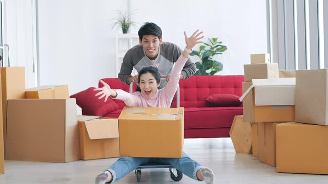 Happy new home Asian couple run push cardboard boxes and look at camera, Couple homeowners playing having fun on moving day celebrate mortgage relocation removals concept.