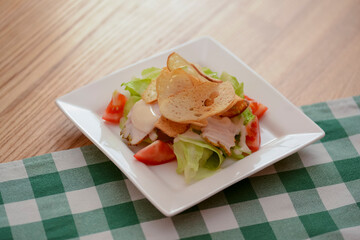 Plate of traditional Caesar Salad with chicken and salad on light wooden table with green plaid tablecloth.