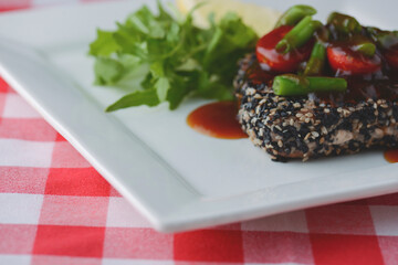 Beef stir-fry steak covered in sesame seeds with vegetables and tomato sauce served on white plate over plaid tablecloth